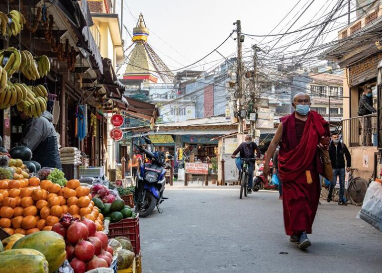 A monk wearing a red robe and a face mask walks down an urban street lined with fruit stalls, a motorcycle, and pedestrians, with a stupa visible in the background and a tangle of electrical wires overhead.