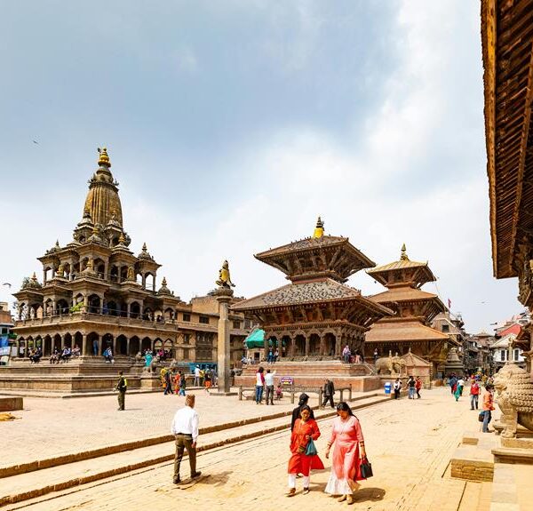 Alt text: A bustling square in a Nepalese city featuring traditional multi-tiered pagoda-style temples and shrines with elaborate wood carvings, bustling with locals and visitors under a cloudy sky.