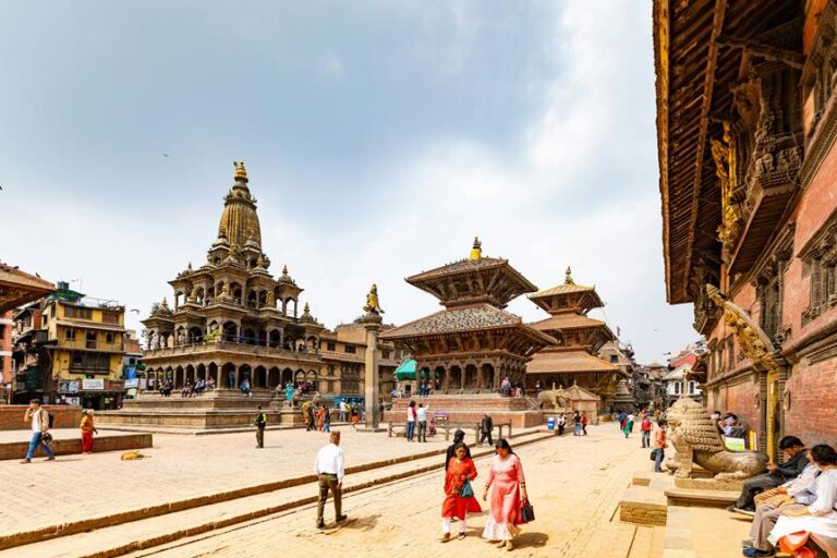 Alt text: A bustling square in a Nepalese city featuring traditional multi-tiered pagoda-style temples and shrines with elaborate wood carvings, bustling with locals and visitors under a cloudy sky.