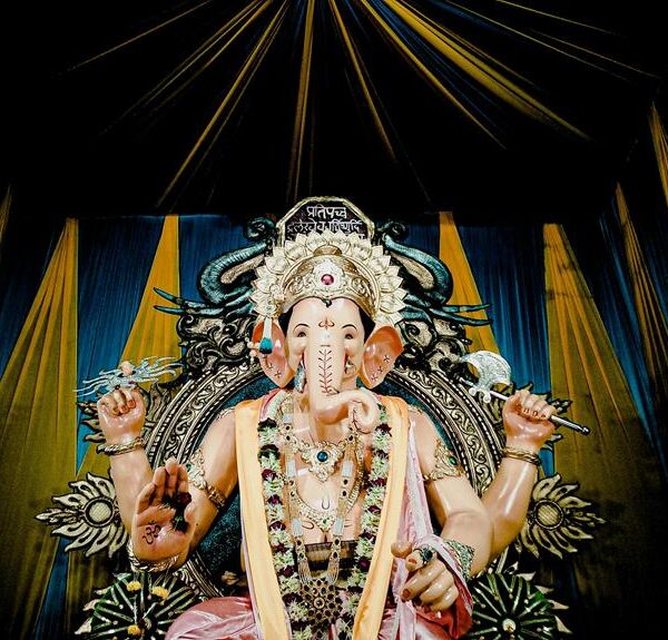 A colorful statue of the Hindu deity Ganesha with a detailed crown and decorations, set against a backdrop with draping blue and gold curtains.