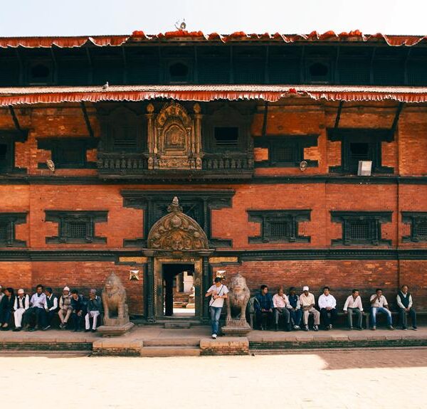 Traditional Nepalese architecture with ornate wood carvings on a multi-tiered pagoda-style building; people sitting and walking in front, under a clear sky.