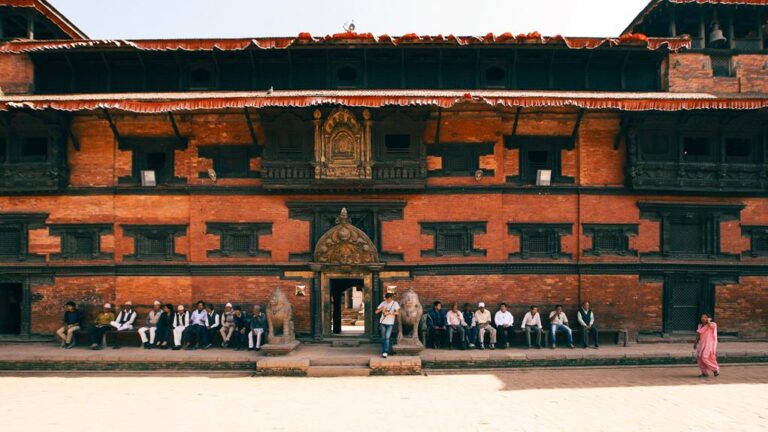 Traditional Nepalese architecture with ornate wood carvings on a multi-tiered pagoda-style building; people sitting and walking in front, under a clear sky.