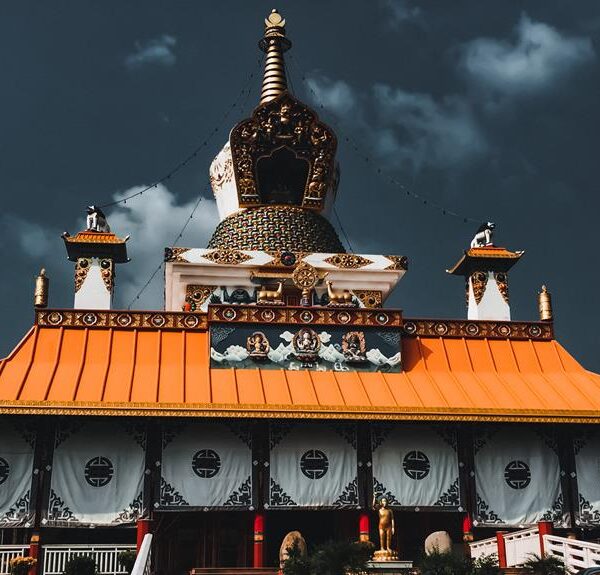 A traditional Tibetan Buddhist temple with a distinct orange roof, intricate decorations, and a multi-colored stupa under a dramatic cloudy sky.