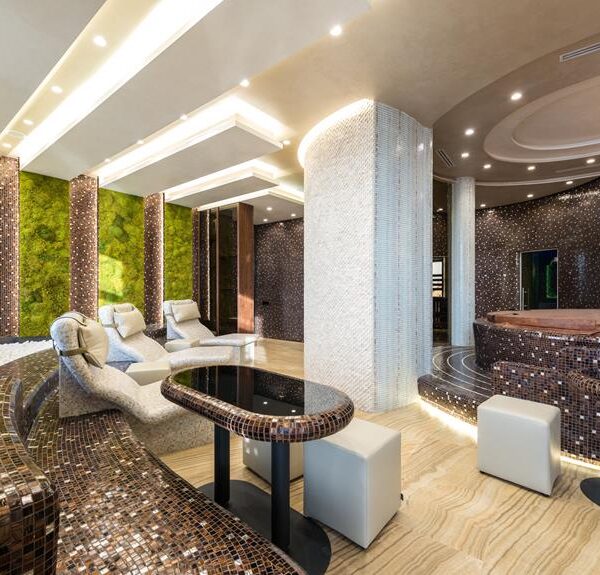 A luxurious interior with moss green walls, patterned brown mosaic columns, a curvaceous mosaic-tiled seating area, modern white chairs, and sophisticated ceiling lighting.
