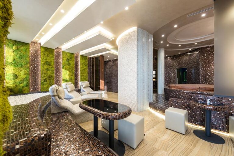 A luxurious interior with moss green walls, patterned brown mosaic columns, a curvaceous mosaic-tiled seating area, modern white chairs, and sophisticated ceiling lighting.