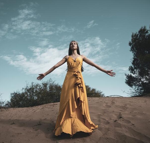 A woman in a flowing yellow dress stands with her arms open and head tilted back on a sandy hill against a blue sky with delicate clouds.