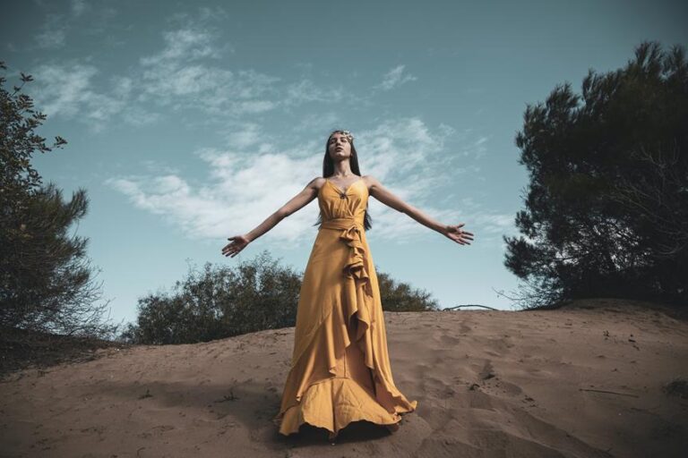 A woman in a flowing yellow dress stands with her arms open and head tilted back on a sandy hill against a blue sky with delicate clouds.