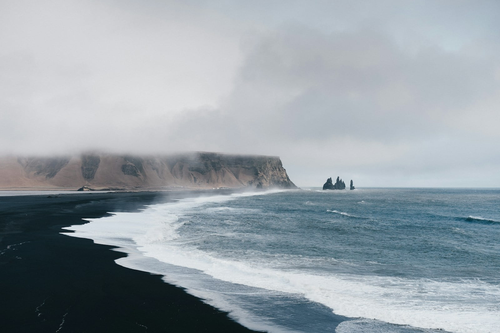 A serene seascape featuring a black sand beach with gentle waves, a misty cliff in the background, and distinctive sea stacks piercing the horizon.