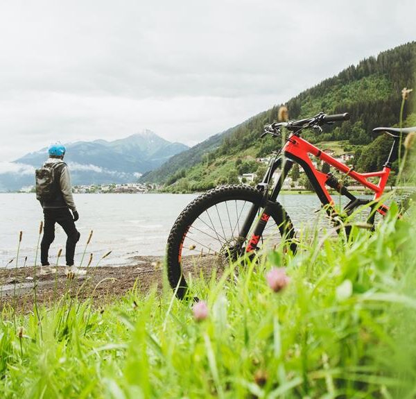 A person wearing a helmet stands by a lake, gazing at the mountains in the distance, with a red mountain bike in the foreground on a grassy shore.