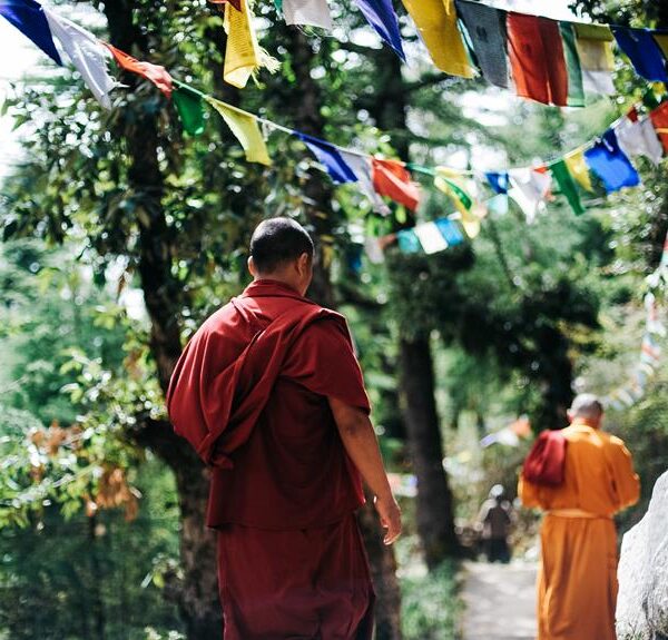 Two monks in traditional red robes walking along a path with colorful prayer flags hanging in the trees.
