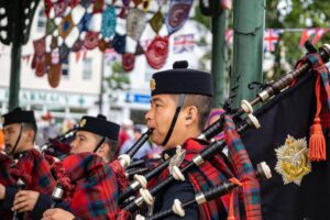 Bagpipers in traditional red tartan kilts and glengarry hats playing in a public setting with British flags in the background.