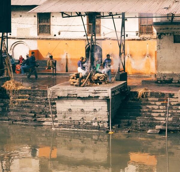 Cremation pyres beside a river with onlookers and mourners gathered around in a ritual setting, reflecting a cultural funeral practice.
