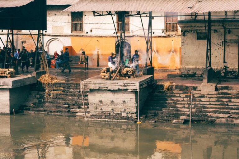 Cremation pyres beside a river with onlookers and mourners gathered around in a ritual setting, reflecting a cultural funeral practice.