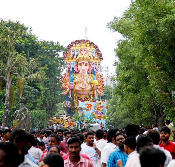 A colorful procession with a large idol of the Hindu deity Ganesha on a decorated platform, surrounded by a crowd of people in a tree-lined street.