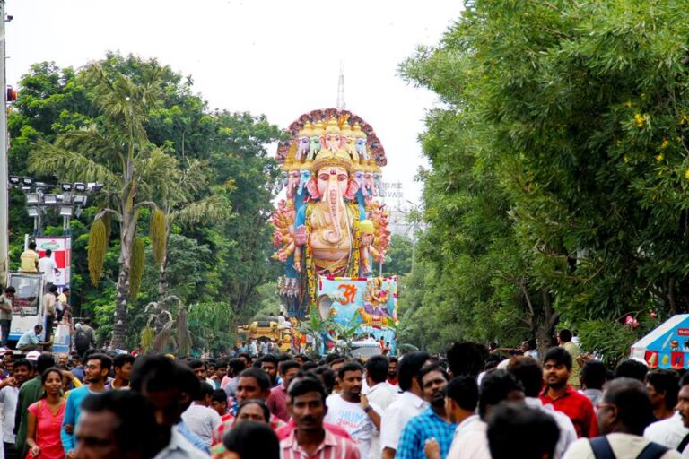 A colorful procession with a large idol of the Hindu deity Ganesha on a decorated platform, surrounded by a crowd of people in a tree-lined street.