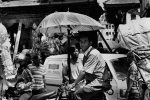 A black and white photo showing a busy street scene with a man riding a bicycle with an umbrella attached to it, multiple people, motorbikes, and a car in the background.