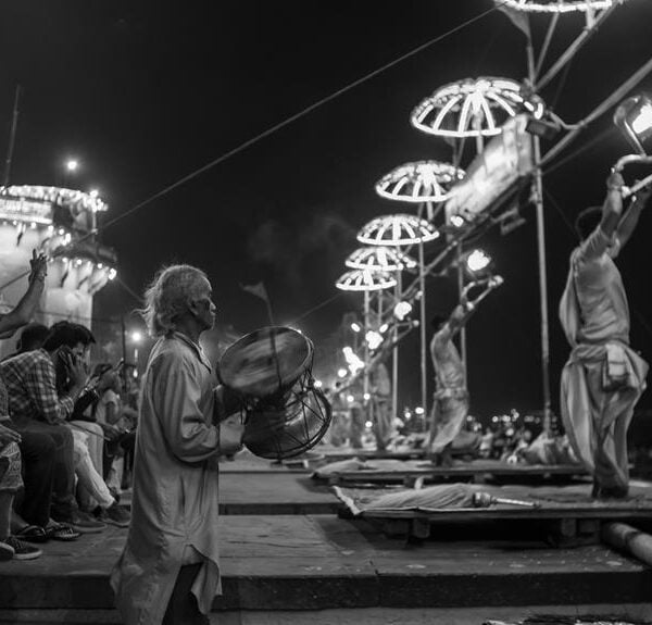 Black and white image of people participating in an outdoor ceremony by a river, with individuals seated on steps, a man playing a drum, and others holding lit traditional lamps on pole structures.