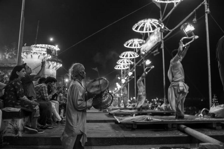 Black and white image of people participating in an outdoor ceremony by a river, with individuals seated on steps, a man playing a drum, and others holding lit traditional lamps on pole structures.
