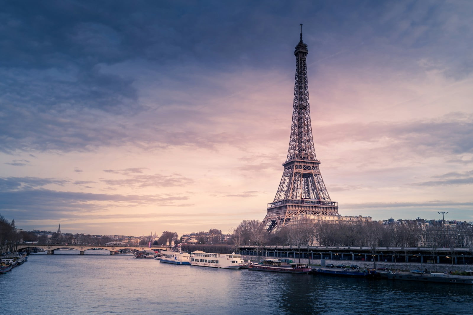 The Eiffel Tower overlooking the Seine River with boats and a bridge against a backdrop of a pastel sunset sky.