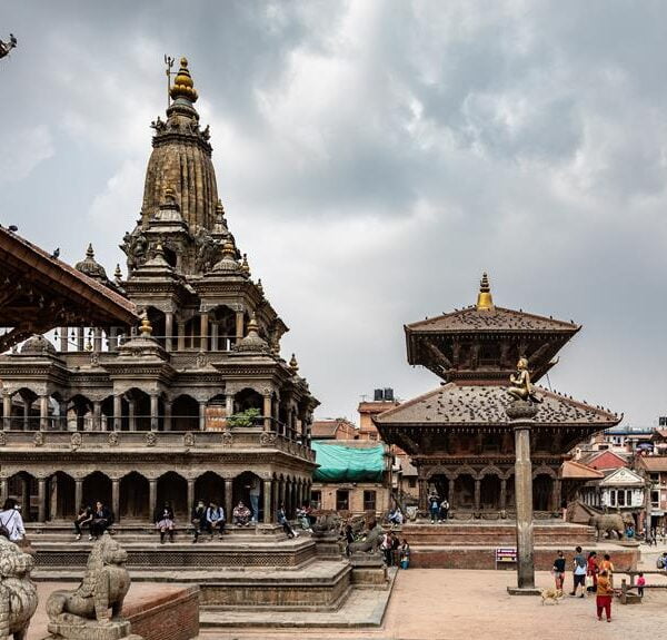 Alt text: A bustling scene at an ancient Nepalese temple complex with intricate architecture, stone statues, and people exploring the historic surroundings under a cloudy sky.