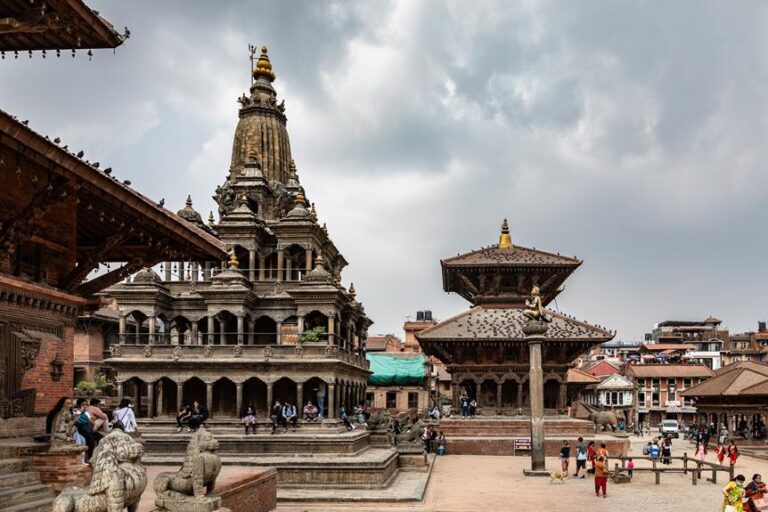 Alt text: A bustling scene at an ancient Nepalese temple complex with intricate architecture, stone statues, and people exploring the historic surroundings under a cloudy sky.