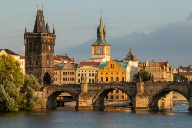 View of the Charles Bridge over the Vltava River during sunset with historical towers and buildings in the background, and people visible on the bridge, in Prague, Czech Republic.