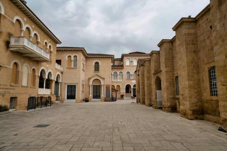 A courtyard with a paved surface flanked by historic buildings with arched windows and doorways under a cloudy sky.