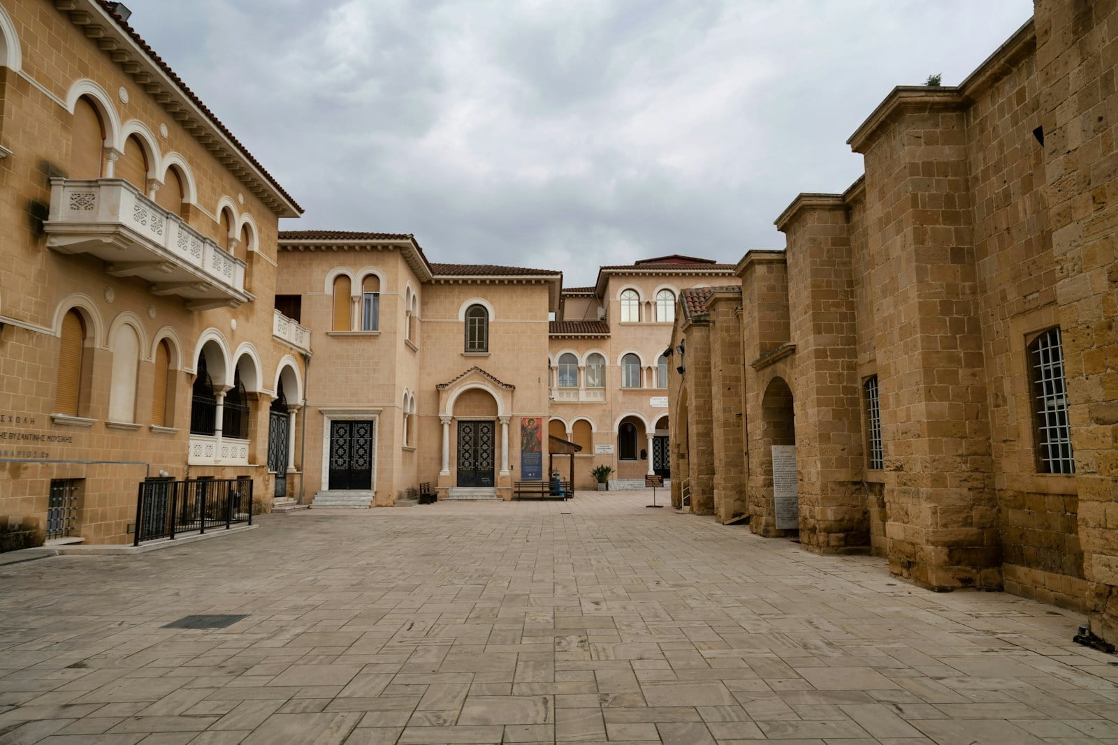 A courtyard with a paved surface flanked by historic buildings with arched windows and doorways under a cloudy sky.