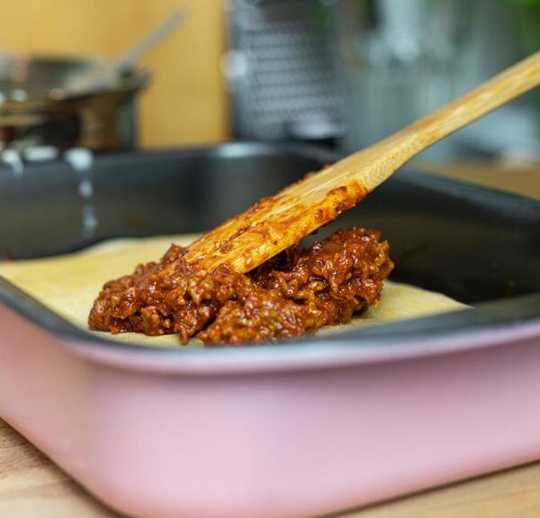 Wooden spoon spreading meat sauce over a layer of pasta in a pink baking dish on a kitchen counter with cookware in the background.