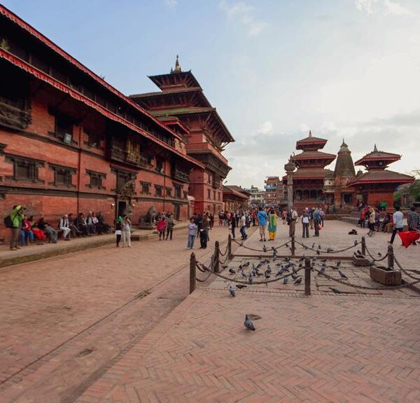 A bustling square with people walking and sitting in front of traditional red brick buildings with tiered pagoda-style roofs, under a partly cloudy sky. Pigeons are scattered on the ground, and a roped-off area with wooden structures is visible in the foreground.