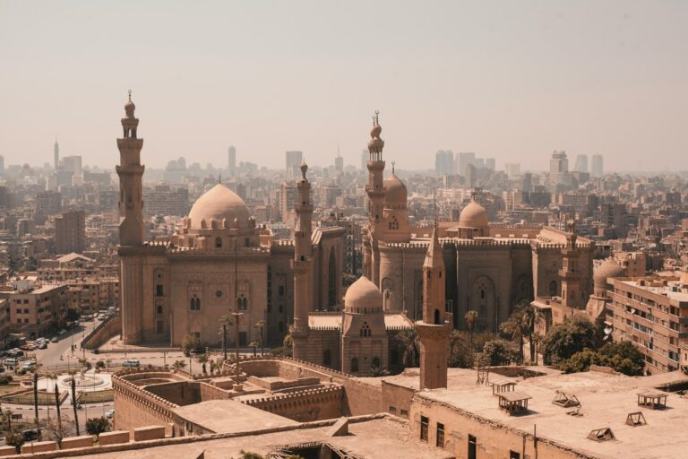 Aerial view of historic Islamic architecture with domes and minarets in Cairo, Egypt, against a backdrop of densely packed modern city buildings under a hazy sky.