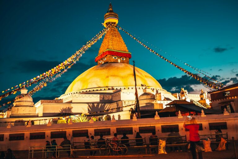 The image depicts Boudhanath Stupa at dusk with illuminated prayer flags extending from the top, vibrant warm lighting on the structure, and people engaging in various activities around its base.