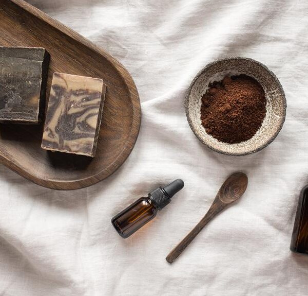 Natural skincare products displayed on a crumpled white linen fabric, including two bars of handmade soap on a wooden tray, a bowl of brown powder with a wooden spoon, and two amber glass bottles, one with a dropper.