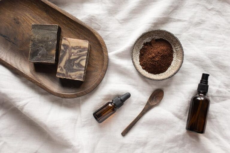 Natural skincare products displayed on a crumpled white linen fabric, including two bars of handmade soap on a wooden tray, a bowl of brown powder with a wooden spoon, and two amber glass bottles, one with a dropper.