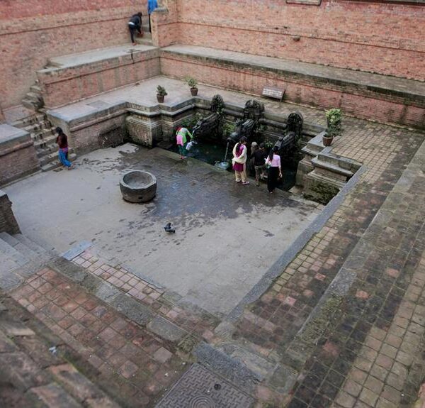 People are gathered around an ancient stone water spout with intricate carvings in a brick-paved courtyard, surrounded by historic walls, with stairs leading down to the spout from one side.