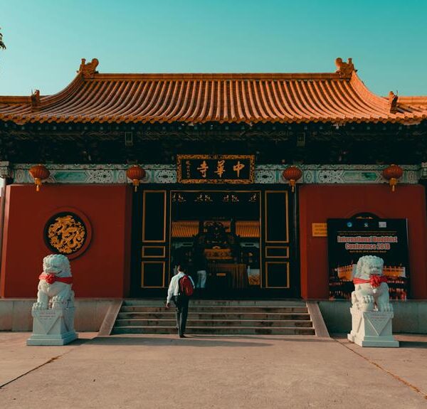 Traditional Chinese temple entrance with red walls, ornate golden Chinese characters, two stone lion statues, and a person walking up the stairs.