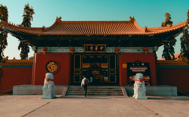 Traditional Chinese temple entrance with red walls, ornate golden Chinese characters, two stone lion statues, and a person walking up the stairs.