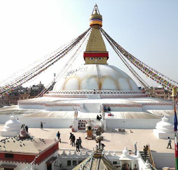 A large white stupa with a golden spire, colorful prayer flags, and visitors walking around the base against a clear sky.