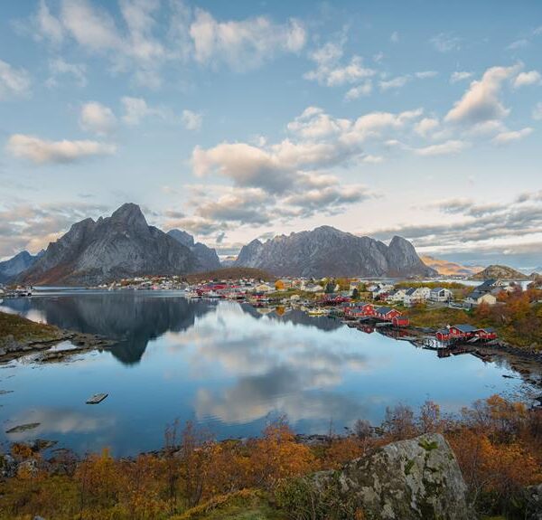 A picturesque view of a colorful village nestled between rugged mountains and a reflective fjord during early evening, with autumn foliage in the foreground and a partly cloudy sky above.