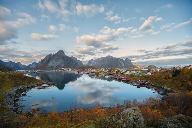 A picturesque view of a colorful village nestled between rugged mountains and a reflective fjord during early evening, with autumn foliage in the foreground and a partly cloudy sky above.