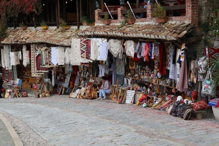 An outdoor market stall with various traditional crafts and textiles on display, set against a backdrop of a cobblestone street and a building with plants and rustic architecture.