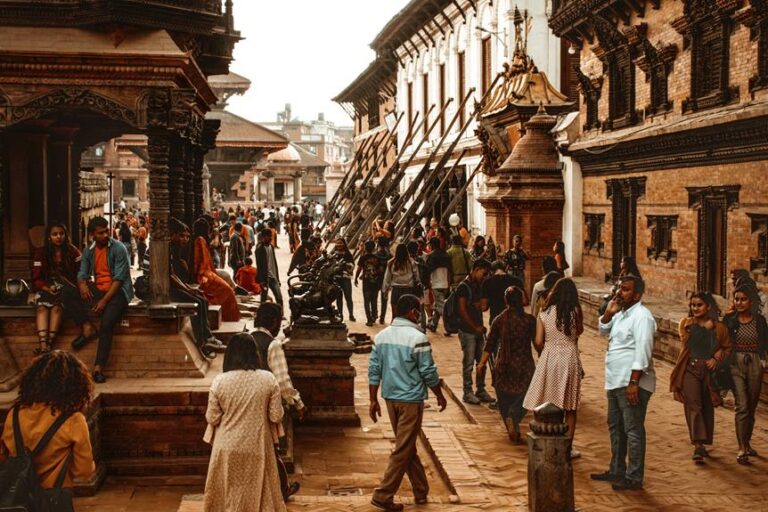 A bustling street scene in an ancient city square with traditional architecture, where people are walking, socializing, and sitting on steps near historic structures and sculptures.