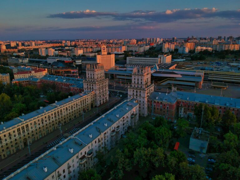 Aerial view of a cityscape during sunset with distinctive buildings illuminated by the warm glow of the setting sun, casting long shadows over the streets and surrounding structures.