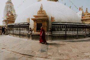 A monk in a red robe walks past a large, white-domed stupa adorned with prayer flags and surrounded by an ornate metal fence.