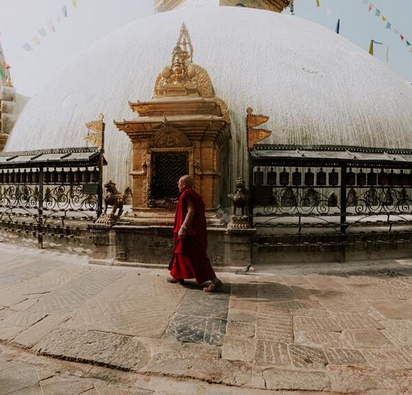 A monk in a red robe walks past a large, white-domed stupa adorned with prayer flags and surrounded by an ornate metal fence.