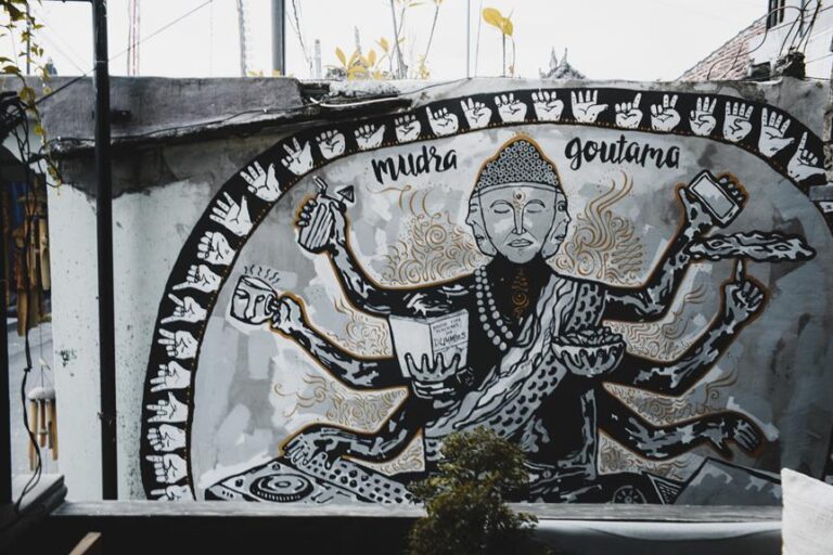 A monochrome mural featuring a stylized representation of a multi-armed figure with text labels "Mudra" and "Goutama". The artwork is adorned with patterns and symbolic imagery, displayed on an urban wall.