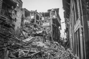 Black and white photo of people navigating through a narrow street filled with debris from heavily damaged and collapsed buildings after a disaster.
