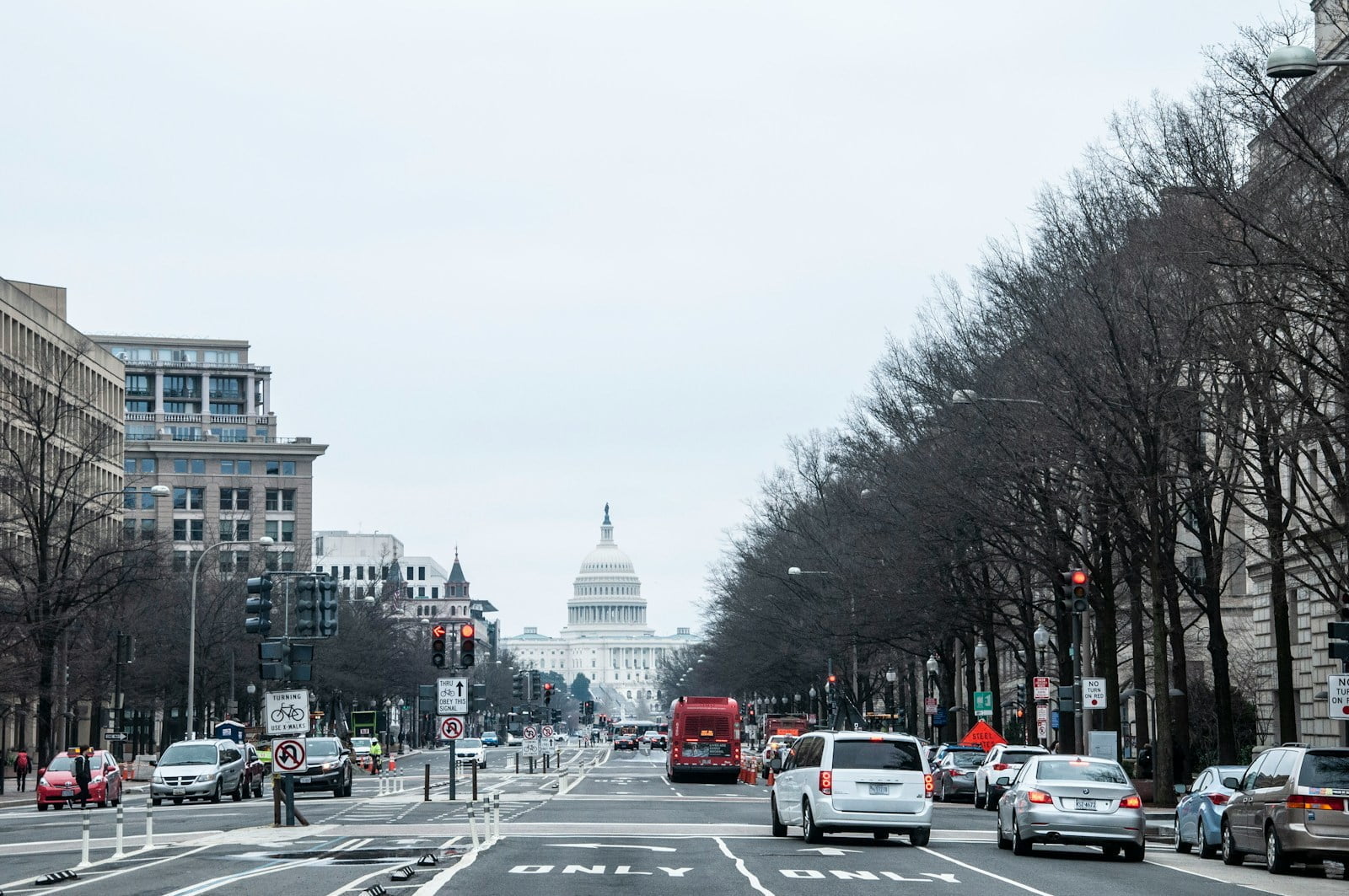 Street view of a busy road leading to the United States Capitol building in the distance, with traffic lights, cars, and leafless trees lining the street under an overcast sky.