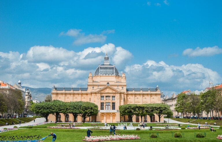 Alt text: A view of an elegant, historic building with a glass dome, surrounded by a green park with blooming flowers, trees, and people enjoying the outdoors under a blue sky with fluffy clouds.