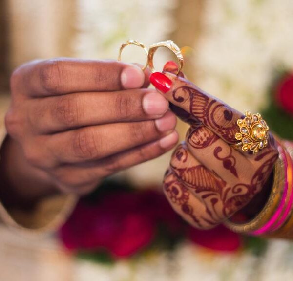 A close-up of a person's hands holding two golden rings, with intricate henna designs on her wrists and forearms, red nail polish, and several gold bangles, set against a background of flowers.
